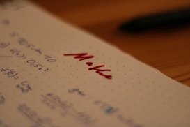 Notebook paper with mathematical equations written in blue ink, alongside the word 'Maths' written prominently in red ink. A pen, placed out of focus, lies on a wooden table in the background.