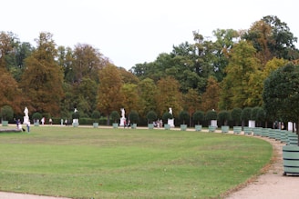 A spacious park landscape features a wide expanse of green lawn bordered by an array of large trees with autumn foliage. Statues are strategically placed along the path, adding a classical touch to the peaceful scene. Several people are walking along the pathways, enjoying the serene environment.