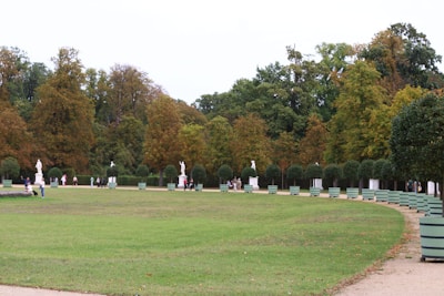A spacious park landscape features a wide expanse of green lawn bordered by an array of large trees with autumn foliage. Statues are strategically placed along the path, adding a classical touch to the peaceful scene. Several people are walking along the pathways, enjoying the serene environment.