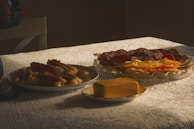 A table is set with a variety of deli meats and cheeses, arranged on different plates. The setup includes cured meats like salami, sliced cheeses, and a large block of cheddar cheese. The lighting casts a warm, soft glow over the lace tablecloth.