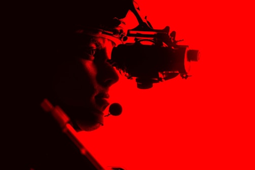 A silhouetted figure is seen side-on against a vibrant red background, wearing night vision goggles. The figure appears to be equipped with a tactical headset, conveying a sense of focus and readiness.