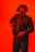 A person wearing a tactical helmet with what appears to be night vision goggles, dressed in a dark suit and tie. The individual is holding a firearm, set against a bright red background, giving the scene a dramatic and intense feel.