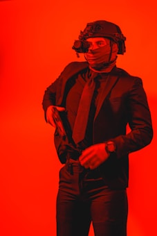 A person wearing a tactical helmet with what appears to be night vision goggles, dressed in a dark suit and tie. The individual is holding a firearm, set against a bright red background, giving the scene a dramatic and intense feel.