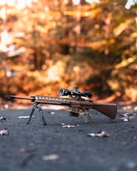A high-powered rifle with a mounted scope and bipod is positioned on a paved surface surrounded by fallen leaves. The background is a blurred, warm, autumnal scene with trees in various shades of orange and brown.
