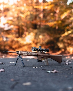 A high-powered rifle with a mounted scope and bipod is positioned on a paved surface surrounded by fallen leaves. The background is a blurred, warm, autumnal scene with trees in various shades of orange and brown.