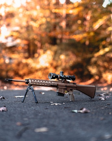 A high-powered rifle with a mounted scope and bipod is positioned on a paved surface surrounded by fallen leaves. The background is a blurred, warm, autumnal scene with trees in various shades of orange and brown.
