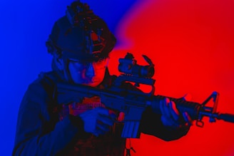 A person in tactical gear and helmet holds a firearm with both hands, set against a vibrant backdrop of blue and red lighting. The firearm features a scope and the person appears focused and alert.