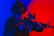 A person in tactical gear and helmet holds a firearm with both hands, set against a vibrant backdrop of blue and red lighting. The firearm features a scope and the person appears focused and alert.