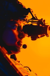 A person is wearing night vision goggles with a microphone in a profile view. The background is brightly lit with an orange hue, creating a vibrant contrast. The equipment and helmet suggest a military or tactical setting.