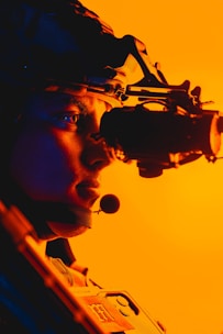 A person is wearing night vision goggles with a microphone in a profile view. The background is brightly lit with an orange hue, creating a vibrant contrast. The equipment and helmet suggest a military or tactical setting.