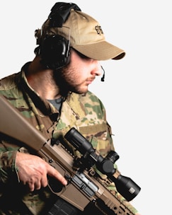 A person wearing camouflage military attire and a beige cap, equipped with over-ear headphones and a microphone. They are holding a tactical rifle with a scope.