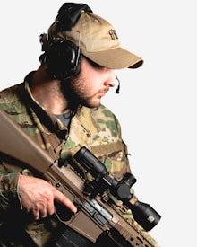 A person wearing camouflage military attire and a beige cap, equipped with over-ear headphones and a microphone. They are holding a tactical rifle with a scope.