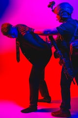 A person in tactical gear and night vision goggles is detaining another person dressed in a suit. The scene is dramatically lit with intense red and blue lighting, creating a high-contrast and tense atmosphere.