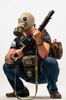 A person is crouching while wearing a gas mask and holding a rifle. They have a backpack and a tactical vest labeled USCG, with additional gear.
