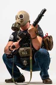 A person is crouching while wearing a gas mask and holding a rifle. They have a backpack and a tactical vest labeled USCG, with additional gear.