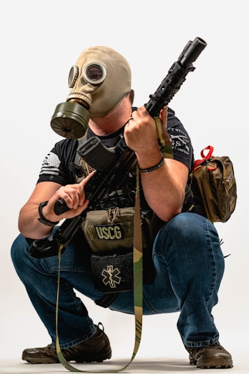 A person is crouching while wearing a gas mask and holding a rifle. They have a backpack and a tactical vest labeled USCG, with additional gear.
