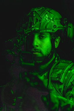 A person wearing military gear, including a helmet with tactical equipment, is illuminated by a green light. The person's face is partially visible with a focused expression.