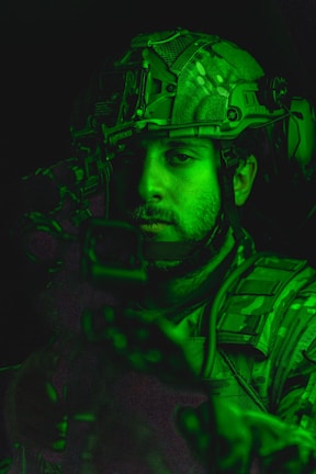 A person wearing military gear, including a helmet with tactical equipment, is illuminated by a green light. The person's face is partially visible with a focused expression.