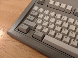 A close-up view of a gray mechanical keyboard with a light cream color scheme. The keyboard keys include letters, numbers, and function keys. It is placed on a wooden surface.