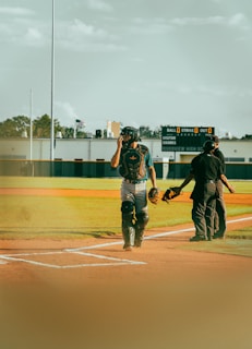 A baseball player wearing catcher's gear stands on a baseball field, adjusting their helmet. An umpire in black attire is present, facing away. The background features a scoreboard displaying zero scores for balls, strikes, and outs. The weather is clear, with a blue sky and some clouds overhead. The setting has a calm, outdoor atmosphere.