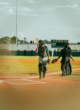 A baseball player wearing catcher's gear stands on a baseball field, adjusting their helmet. An umpire in black attire is present, facing away. The background features a scoreboard displaying zero scores for balls, strikes, and outs. The weather is clear, with a blue sky and some clouds overhead. The setting has a calm, outdoor atmosphere.