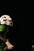 A football player wearing a green uniform and a white helmet is holding a football. The background is dark, emphasizing the player who is partially lit. The player's helmet is prominently displayed with a number and logo.