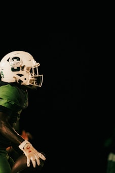 A football player wearing a green uniform and a white helmet is holding a football. The background is dark, emphasizing the player who is partially lit. The player's helmet is prominently displayed with a number and logo.