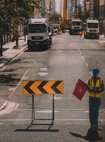 A city street is under construction with multiple trucks parked and a crane visible in the distance. A road sign with black arrows on a yellow background indicates a detour or direction change. A construction worker wearing a hard hat and a safety vest is holding a red flag.