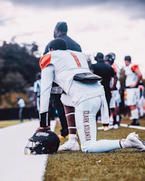 A football player wearing a white uniform with orange accents is kneeling on the grass next to a sideline, holding a black helmet with 'CAU' lettering. Other players and coaches are visible in the background.