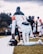 A football player wearing a white uniform with orange accents is kneeling on the grass next to a sideline, holding a black helmet with 'CAU' lettering. Other players and coaches are visible in the background.