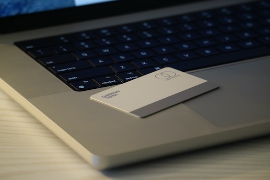 A close-up of a laptop keyboard with a card placed on the touchpad. The card appears to have a simple design with some text and a logo. The laptop is on a wooden surface, and the lighting creates a soft, muted ambiance.