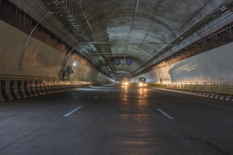 A dimly lit tunnel with cars driving inside. The tunnel features smooth, curved walls and a high ceiling with exposed infrastructure such as cables and pipes. Yellow and white lines are painted on the road surface for lane guidance.
