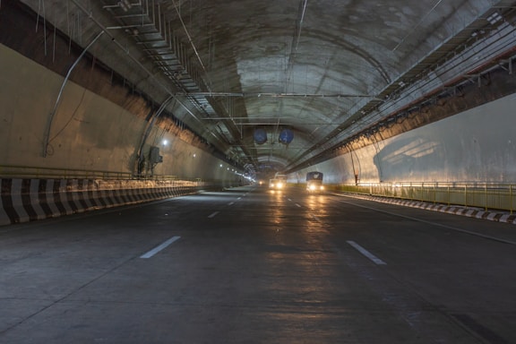 A dimly lit tunnel with cars driving inside. The tunnel features smooth, curved walls and a high ceiling with exposed infrastructure such as cables and pipes. Yellow and white lines are painted on the road surface for lane guidance.