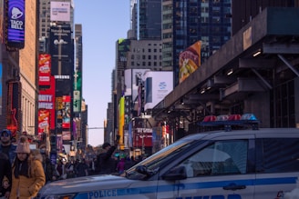 A busy urban street scene filled with a crowd of people, bright billboards, and tall skyscrapers, suggesting an iconic city atmosphere. A police car is parked in the foreground. The setting is lively and bustling with advertisements for various brands visible.