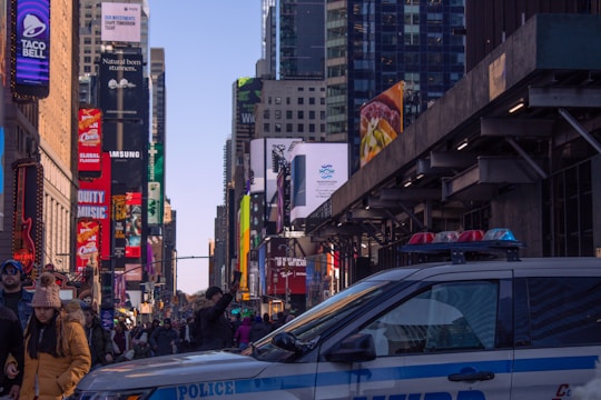 A busy urban street scene filled with a crowd of people, bright billboards, and tall skyscrapers, suggesting an iconic city atmosphere. A police car is parked in the foreground. The setting is lively and bustling with advertisements for various brands visible.