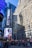 A bustling urban scene features tall skyscrapers with various architectural designs, including a brick building with many windows and a modern glass structure. The area is filled with large, colorful digital billboards, one of which displays an advertisement for a luxury brand. The street below is crowded with people, and there are food stalls with yellow and blue umbrellas. The sunlight casts dramatic shadows on the buildings, highlighting the vibrant atmosphere.