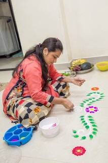 A woman is engaged in creating a colorful rangoli design on the floor. She is applying white powder and various colored powders to form intricate patterns, including flowers and leaves. She is dressed in a traditional outfit with a coral top and a multi-colored skirt. In the background, there are various fruits and items placed in bowls on the floor.