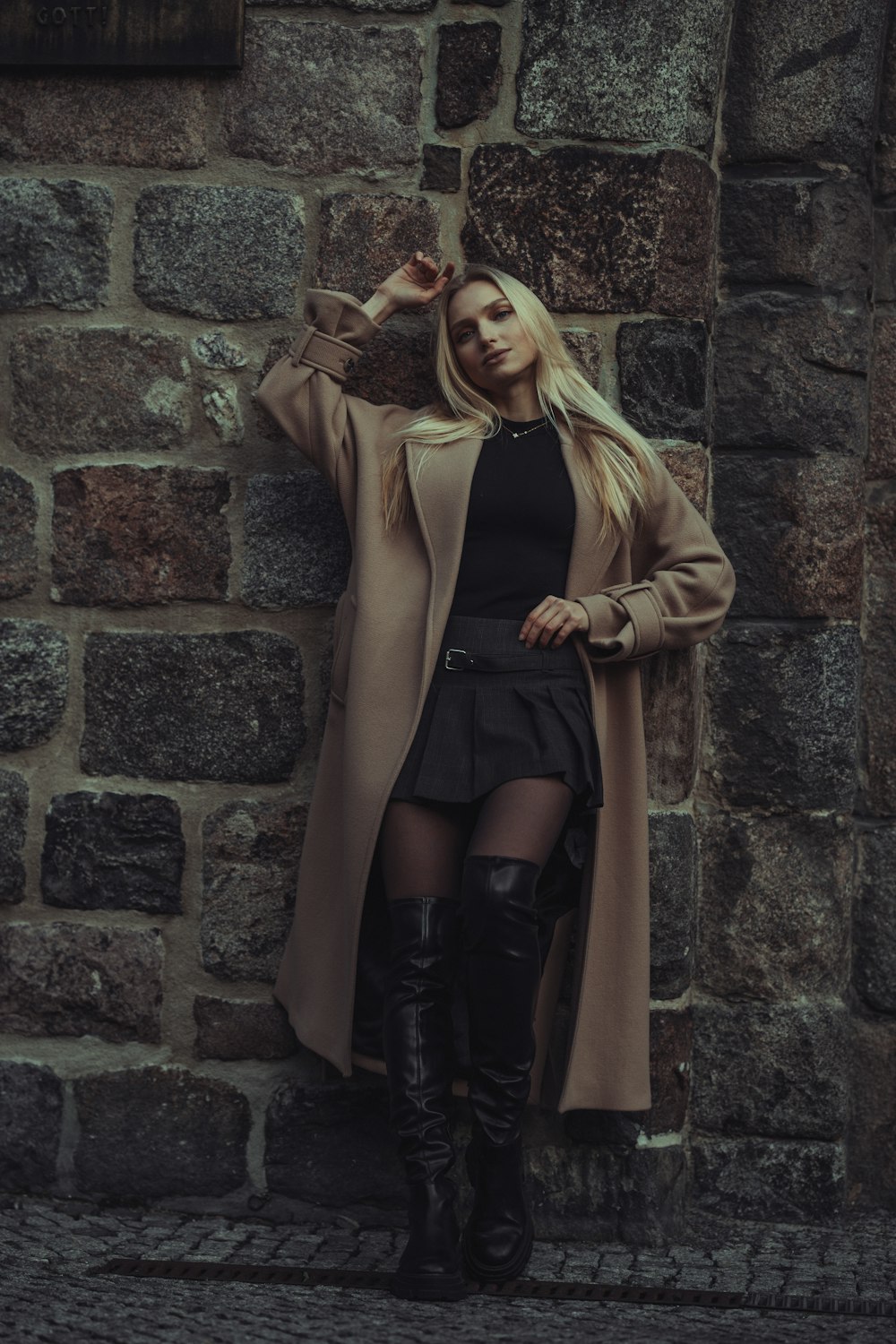 a woman in a black dress and boots leaning against a brick wall