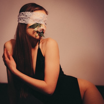 A person with long hair is partially reclining while wearing a lace blindfold. There are flowers and leaves placed across the lower part of the face. The person is dressed in a black outfit, and the overall setting has a soft, warm-toned lighting.