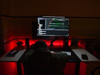 A person is sitting at a desk in a dimly lit room, facing a large computer monitor displaying code. The workspace is illuminated by red and blue lights, adding a moody atmosphere. Various items are scattered on the desk, including notebooks, a phone, and a small picture frame.