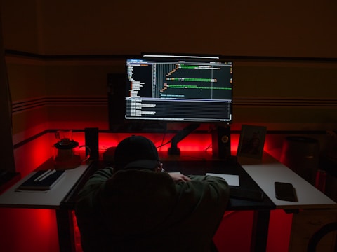 A person is sitting at a desk in a dimly lit room, facing a large computer monitor displaying code. The workspace is illuminated by red and blue lights, adding a moody atmosphere. Various items are scattered on the desk, including notebooks, a phone, and a small picture frame.