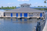 A building with gray siding and a dark gray roof sits on a body of water, resembling a floating dock structure. It features several blue doors and has various signs, including one that indicates firewood is for sale. In the background, there is greenery, with trees and a clear blue sky, and a walkway leading to the building.