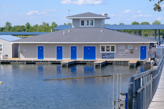 A building with gray siding and a dark gray roof sits on a body of water, resembling a floating dock structure. It features several blue doors and has various signs, including one that indicates firewood is for sale. In the background, there is greenery, with trees and a clear blue sky, and a walkway leading to the building.