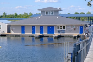 A building with gray siding and a dark gray roof sits on a body of water, resembling a floating dock structure. It features several blue doors and has various signs, including one that indicates firewood is for sale. In the background, there is greenery, with trees and a clear blue sky, and a walkway leading to the building.