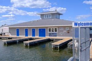 A waterfront building features multiple blue doors and a gray roof with a central tower. Wooden walkways extend over a body of water surrounding the structure. Signs on the building advertise services and products, including a notice that they are open daily and firewood for sale. The sky above is bright with scattered clouds.