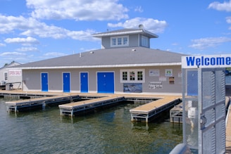 A waterfront building features multiple blue doors and a gray roof with a central tower. Wooden walkways extend over a body of water surrounding the structure. Signs on the building advertise services and products, including a notice that they are open daily and firewood for sale. The sky above is bright with scattered clouds.