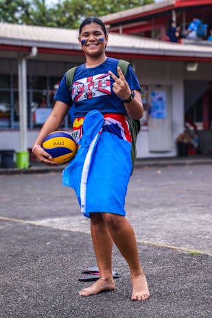 A person smiling and making a peace sign while holding a volleyball. They are dressed in a t-shirt, traditional skirt-like attire in blue with colorful patterns, and carrying a backpack. The background shows an outdoor area near a building with a visible roof and people in the back.