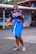 A person smiling and making a peace sign while holding a volleyball. They are dressed in a t-shirt, traditional skirt-like attire in blue with colorful patterns, and carrying a backpack. The background shows an outdoor area near a building with a visible roof and people in the back.