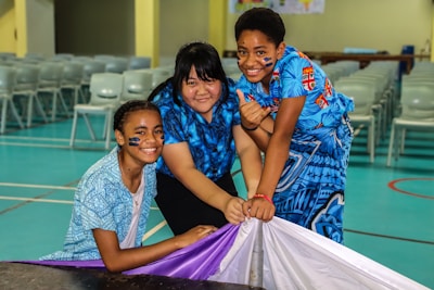 Three children wearing colorful blue shirts are smiling and kneeling on a gym floor, holding a purple and white cloth. They have face paint, indicating participation in a festive or sporting event. The background features a row of empty chairs.