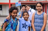 A diverse group of five smiling children standing outdoors near a building with red and white walls. They are wearing colorful clothing, and one child is holding a small flag.